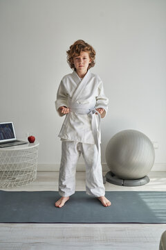 Focused young boy with curly hair in white kimono standing on mat in front of laptop at home looking at camera while learning hands position in judo