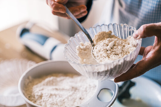 From above unrecognizable confectioner in apron standing in kitchen with glass bowl and adding flour with spoon while preparing dough for muffins
