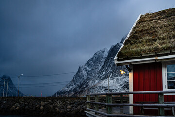 Cottage with red striped walls and white window frames on wooden pier on strait shore against snowy township at foothill in winter cloudy day in Lofoten