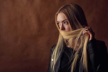 Beautiful young girl in black leather covers her face with hair on a brown background. Studio photography. Space for text