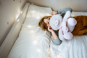 Excited little girl hugging plush toy and laughing while lying on soft bed near fairy lights at home
