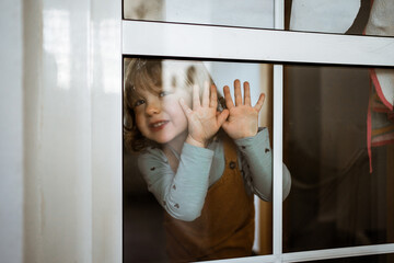 Cute little girl in casual clothes looking at camera and smiling while standing behind window at home and touching glass