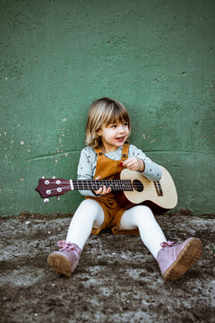 Little girl with ukulele sitting on rough ground near kick scooter against weathered green wall on street