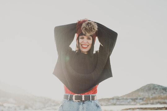 From below of happy active young female dressed in hat with fur pompom and sweater with jeans having fun and looking at camera enjoying nice sunny winter day at countryside