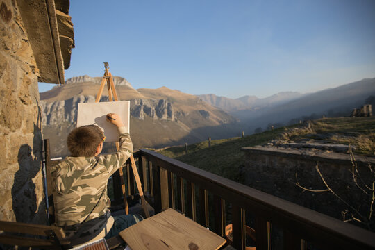 Back view of faceless boy sitting at easel on picturesque veranda of country house and drawing picture in Cantabria