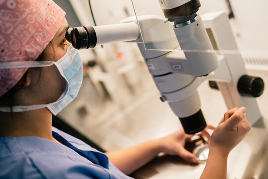 From above doctor in medical mask and uniform injecting ovum on Petri dish and examining cell through microscope in laboratory of modern fertility clinic