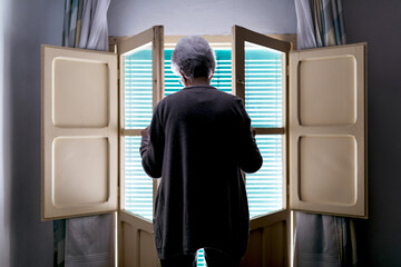 Back view of unrecognizable senior female opening window with shutters while standing in room at home and spending day alone during self isolation because of coronavirus outbreak