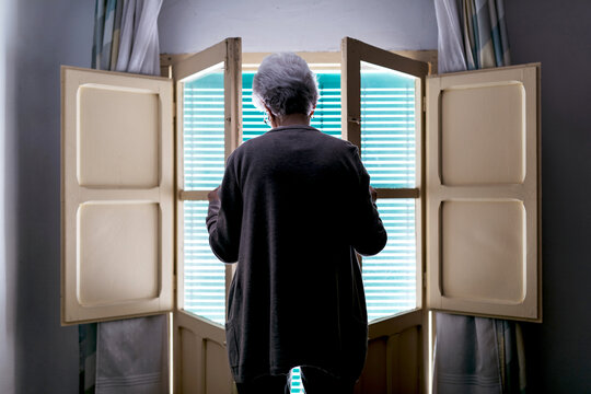 Back view of unrecognizable senior female opening window with shutters while standing in room at home and spending day alone during self isolation because of coronavirus outbreak