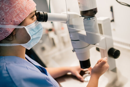 From above doctor in medical mask and uniform injecting ovum on Petri dish and examining cell through microscope in laboratory of modern fertility clinic