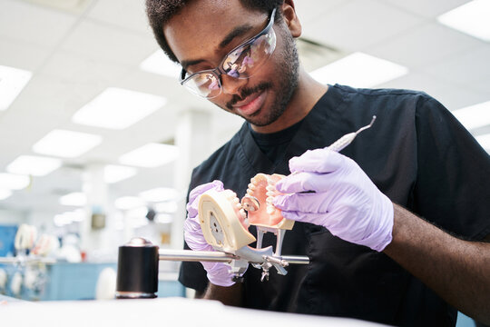 African American guy in latex gloves using mouth mirror and probe to check false teeth while working in modern laboratory
