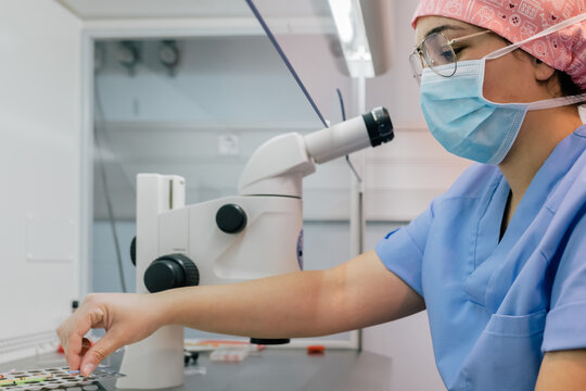 Side view of woman in medical uniform and mask using modern microscope to examine human cells while working in lab of modern clinic