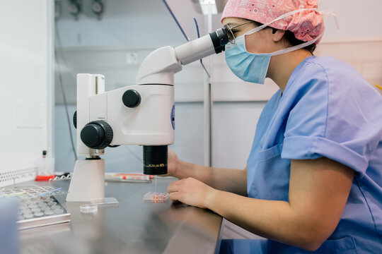 Side View Of Woman In Medical Uniform And Mask Using Modern Microscope To Examine Human Cells While Working In Lab Of Modern Clinic