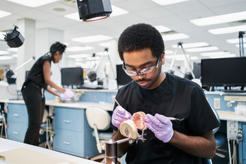 African American guy in latex gloves using mouth mirror and probe to check false teeth while working in modern laboratory