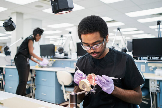 African American guy in latex gloves using mouth mirror and probe to check false teeth while working in modern laboratory