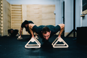 Strong sportsmen in active wear using push up handles during intense training in spacious gym