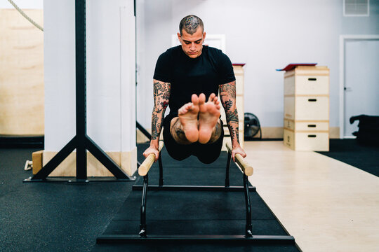 Tattooed strong male athlete doing abdominal exercises using parallel bars during training in gym