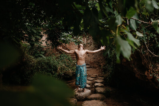 Full body unrecognizable muscular shirtless male in traditional colorful pants meditating while standing on narrow stone pathway among green trees