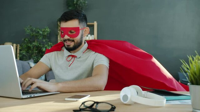 Portrait of young man in superhero costume working with laptop in apartment typing sitting at desk alone and smiling. Modern technology and occupation concept.