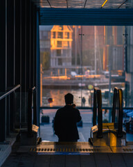 Man on a phone heading down an escalator with city buildings in the distance. The sun is setting.