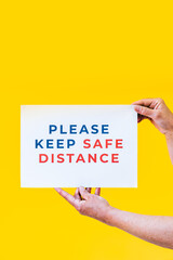 Please keep safe distance. Hands holding a sign for people to respect social distancing measures with an orange background.