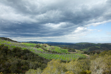 Fototapeta na wymiar Dramatic sky with blue, gray clouds over green valley and hill sides covered with orchard