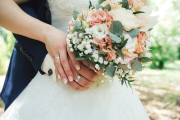 Newlyweds holding colorful wedding bouquet against white wedding gown. Close up of hands with marriage rings on fingers.Outdoor background.wedding day