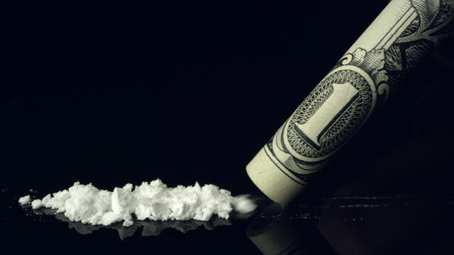 Line of cocaine being snorted up rolled up dollar bill on black reflective table top.