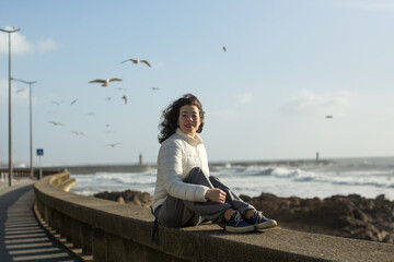 Fototapeta premium Mixed race woman sitting of sea promenade in Porto, Portugal. Seagulls fly over the foaming surf.