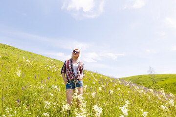 Happy adventurer female stands on the green mountain slope among flowering pink rhododendrons and looking into the distance. Epic travel in the mountains. Wide angle.