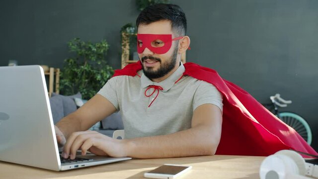Slow motion of young freelancer in superman costume typing with laptop at home working with computer, red cape is moving in wind. People and success concept.