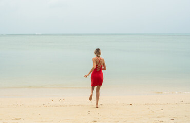 Fototapeta na wymiar Young woman in a red dress runs and jumps on a white sand beach into tropical sea. Young happy woman running on the beach. Happiness to be on a sea or ocean. Vacation and travel concept.