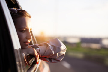 Woman riding on car as passenger looking out of window, enjoy summer, freedom, weekend, wind and hot weather, with sunlight of morning or sunset, dreaming and smiling