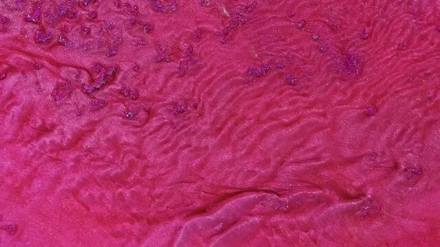 A stream of pink liquid paint with particles of silver sparkles. It recalls the movement of living cells inside an organism or flowing rivers from a bird's eye view.