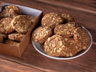 handmade oatmeal cookies on a white plate and many cookies in a cardboard box in the background on a wood