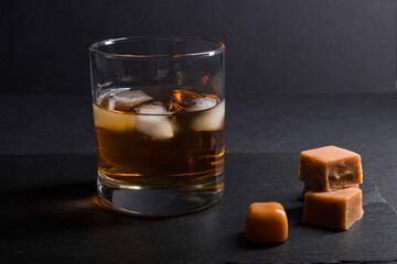 Glass of whiskey with ice and caramel candies on a black stone slate board on black background. Side view, low key, copy space.