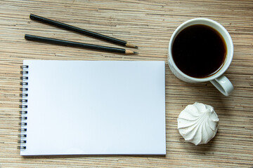 A cup of coffee with marshmallow, notebook with two pencils on wooden background. Cosy workspace. Top view, flat lay