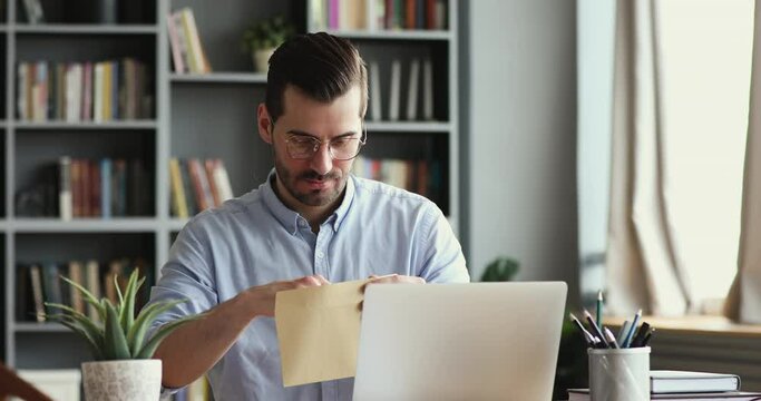 Curious smiling young man in glasses opening paper envelope, reading letter with good news. Happy handsome businessman received investment notification or banking loan approval or salary bonus.