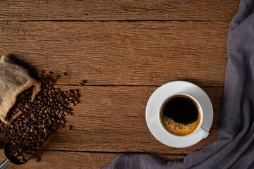 Top view above of Hot fresh black coffee with milk foam in a white ceramic cup with coffee beans roasted in sack bag on wood table background. Flat lay with copy space.