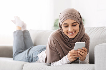 Beautiful arabic girl in hijab lying on couch and texting on smartphone