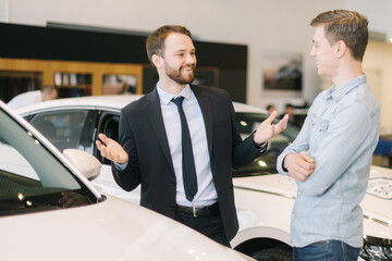 Professional car salesman wearing business suit is telling interested buyer about car in auto...
