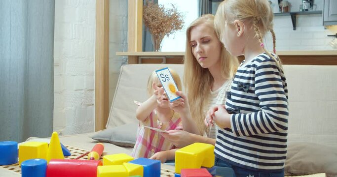 Young mother showing two daughters cards with letters and pictures studying the English alphabet while sitting on the sofa at home. Preschool education.
