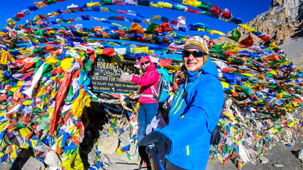 A couple standing between prayer flags at the top of Thorung La Pass, Annapurna Circuit Trek, Nepal. Memorial board. Colorful prayer flags attached to the stone wall, blown by the wind. Clear sky.