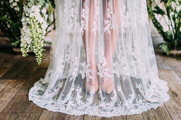 Womans legs with beige shoes in a transparent lace wedding dress and green flowers