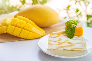 A mango crepe cake sliced on a white plate decorated with a piece of mango fruits and peppermint on a wite table with mango fruit and green nature background.