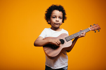 Portrait of a handsome dark-skinned boy playing on a ukulele and looking at the camera in the studio on an orange background.