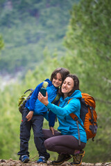 Naklejka na ściany i meble A child with a backpack takes a selfie on a smartphone with mom on the background of a mountain forest.