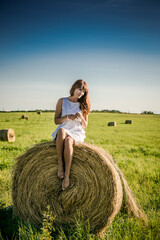 Naklejka na ściany i meble Girl in a white dress on a mown wheat field. Sheaves of hay on the field. girl on the field with hay. Woman near a sheaf of hay in a field. Rural life. Holidays in the village