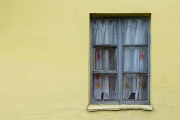 Obraz na płótnie Canvas One old wooden window with peeling paint, with floral curtains. Plastered yellow lemon facade of a poor private house.