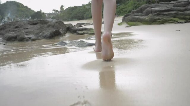 Close Up Young Girl's Legs Walking Barefoot On The Beach With Sea Water Splashing On The Sand. Low Angle Rear View Of Relaxing Female Teenager Walks Along The Coast. Follow Shot And Slow Motion. 