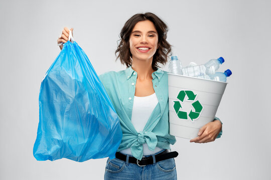 recycling, waste sorting and sustainability concept - smiling young woman in striped t-shirt holding rubbish bin with plastic bottles and trash bag over grey background
